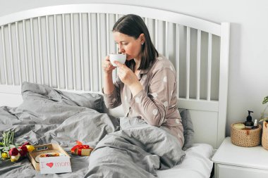 Anneler Günü 'nde yatakta kahvaltı. Pijamalı, kahve içen genç bir kadın. Hediyeler, laleler ve çocukların kartpostalları..