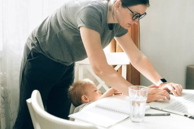 Anne, çocuğu masanın yanında dururken evde dizüstü bilgisayarla çalışmaya çalışıyor. Çocuk bakımı ve çalışan anne kavramı. Bebekle evde çalışan bir kadın..