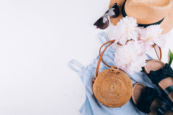 Feminine summer accessories: dress, rattan bag, straw hat, black sandals peony flowers on white background. Summer background. Flat lay, top view.
