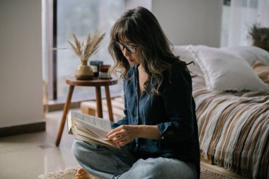 Genç odaklı Asyalı kadın gözlük takarak kitap okurken Lotus 'ta poz verirken rahat oturma odasında kuru buğday ve mumlarla süslenmiş, boş zamanlarını evde geçiriyor.