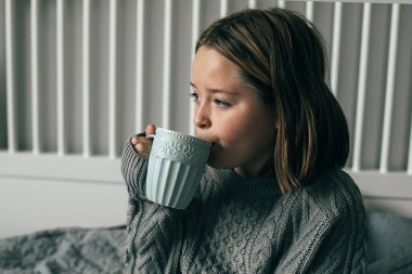 Gri örgü kazaklı, yatak odasındaki rahat yatakta sıcak çikolata içen 8-9 yaşlarında genç bir kız. Sonbahar ya da kış hafta sonu kavramı