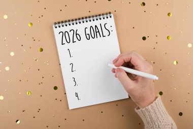 A hand is writing on a blank notepad labeled 2026 Goals, with golden confetti scattered around, representing motivation and future aspirations for personal growth