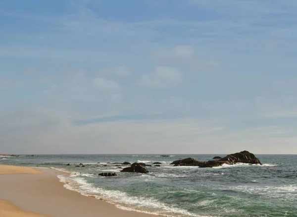 A tranquil coastal scene featuring waves crashing gently, sandy beach with scattered rocks in the ocean. The bright blue sky and untouched shoreline create a peaceful and natural atmosphere, ideal for themes of travel, relaxation, and scenic nature