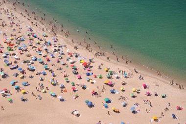 Yukarıdan bir sürü insanın farklı şeyler yaptığı kumsal görünüyor. Açık şemsiyeli renkli atmosfer, şeffaf sular.