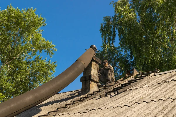 Chimney sweeper cleaning brick chimney while sitting on old building roof in summer day. Rauchfangkehrer at work