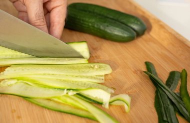 Ahşap pişirme tahtasına büyük bir bıçakla salatalık dilimlemek. Uzun ince salatalık şeritlerinden taze sebze salatası yapan bir kadın evde yemek pişirir.