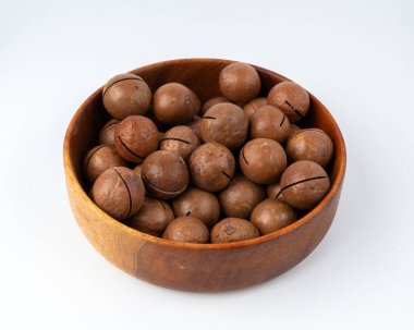 Ahşap kasedeki Macadamia fındıkları izole edilmiş. Doğal soyulmuş macadamia tohumları ve kabukları, beyaz arka planda dünyanın en sert ve en pahalı fındıkları.
