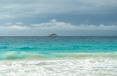 Tropik turkuaz okyanus dalgaları Seyşeller 'deki kumlu sahilde kırılıyor bulutlu gökyüzünün altında uzak bir ada doğal güzelliği, yaz cennetini ve huzurlu kıyı manzarasını yakalıyor.