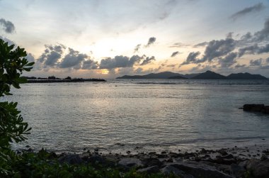 Praslin Adası 'nın silueti ve alçak gelgitte ön planda bitkilerle La Digue' den akşam manzarası