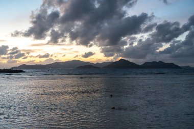 Gün batımında La Digue 'den Praslin Adası manzarası katmanlı tepeler ve ön planda hafif dalgalarla