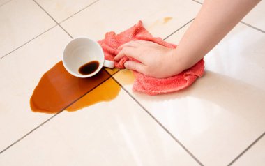 Dökülmüş kahve, kadın fayanslı zemine kahve lekesini siliyor, beyaz bardakla karışık sızıntı, kahverengi içki dökülmüş temizlik.