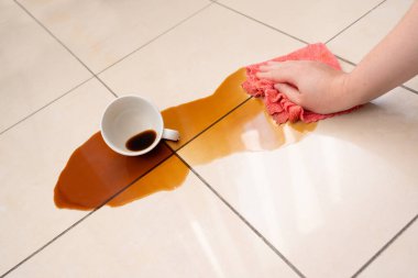 Dökülmüş kahve, kadın fayanslı zemine kahve lekesini siliyor, beyaz bardakla karışık sızıntı, kahverengi içki dökülmüş temizlik.