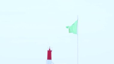 Yeşil güvenlik bayrağı, arka planda kırmızı deniz feneri olan bir sahilde güvenli koşulları gösteriyor.