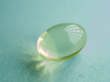 Deniz mavisi yüzeyinde dinlenen tek bir saydam sarı-yeşil softgel hapının makro görüntüsü, sağlık, takviye, beslenme ve ışıltılı yansıma ve yumuşak ışıklandırma ile minimum ilaç tasarımı..