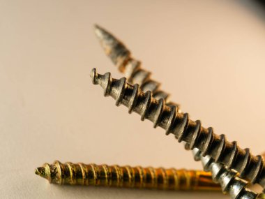 Yumuşak bej bir yüzeye üç vida çakılmış. Bir altın, iki çelik, iplikleri keskin bir şekilde tanımlanmış. Işık kenarları yakalar, derinlik ve kontrastı yaratır. Görüntü araç gereç ve işçilik öneriyor.