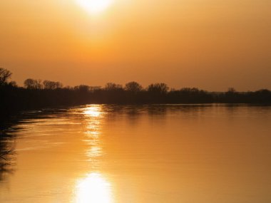 Altın güneş sakin nehir yüzeyine doğru batar. Sessiz sular sıcak, parlayan ışığı yansıtır. Ağaçlar sisli ufukta karanlık duruyor. Gün geçtikçe durgunluk anı tutar. Sakin sahneler için ideal