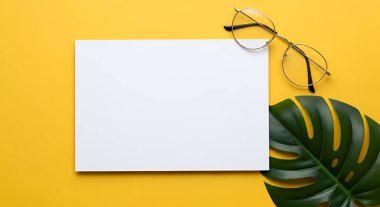 Sarı zemin üzerine boş beyaz bir kağıt yerleştirilir, yanında gözlük ve canavar yaprağı ile, canlı ve davetkar bir sahne yaratır..