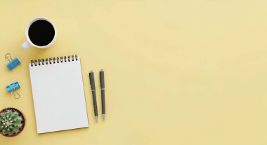 Bir fincan kahve, boş bir defter, kalemler, klipler ve sarı arka planda küçük sulu bir bitkinin yer aldığı minimalist bir çalışma alanının genel görüntüsü..