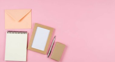Bir defter, resim çerçevesi ve kırtasiye malzemelerinin yukarıdan çekimleri yumuşak pembe bir arkaplanda, temiz ve düzenli bir çalışma alanı estetiği yaratıyor..