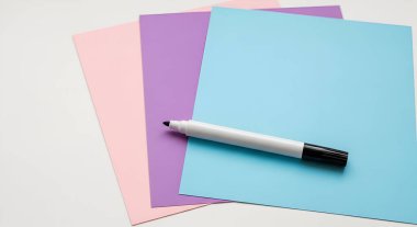 Beyaz bir yüzeye istiflenmiş pembe, mor ve mavi kağıt çarşafların yukarıdan aşağı görüntüsü, mavi sayfada duran siyah bir işaret..