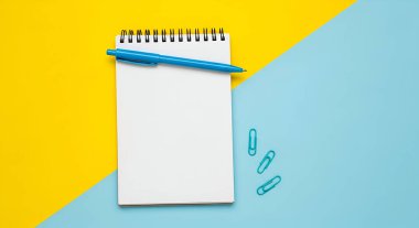 Mavi kalem ve ataçlı boş bir not defteri sarı ve mavi arkaplan üzerine yerleştirilir, notlar için mükemmeldir..