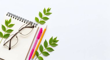 Beyaz arka planda bir defter, gözlük, kalem ve yeşil yaprakların yer aldığı temiz, minimalist bir çalışma alanı yeni ve düzenli bir estetik yaratıyor..