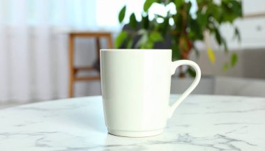 Beyaz bir kupa mermer bir masada oturur, bir bitki ve bulanık arka plan ile sahne derinlik ekleyerek.