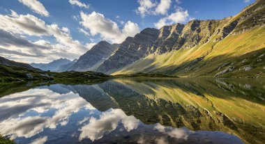 Sakin bir dağ gölü parlak bir gökyüzünün altında görkemli tepeleri yansıtır. Sahne huzurlu, yansıma mükemmel, nefes kesici doğal güzelliği yakalıyor..