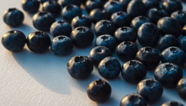 Beyaz bir yüzeye dağılmış taze yaban mersinlerinin canlı renklerini ve dokularını gösteren yakın plan bir görüntüsü. Görüntü, meyvenin doğal güzelliğini vurgular..