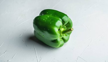 Canlı yeşil bir dolma biber, beyaz dokulu bir yüzeye oturur ve yakın plan çekimde taze, gevrek görünümünü sergiler. Biberler pürüzsüz cilt ışığı yansıtır.