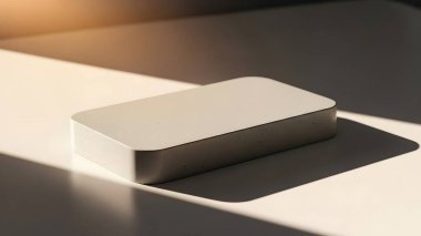 Güneş ışığıyla aydınlatılmış, güçlü bir gölge yaratan basit bir beyaz kutu yüzeyde duruyor. Olay yeri temiz ve minimal..