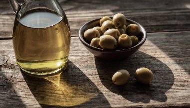 Altın zeytinyağıyla dolu cam bir sürahi yıpranmış ahşap bir yüzeyde taze yeşil zeytinlerle dolu bir kâsenin yanında duruyor..