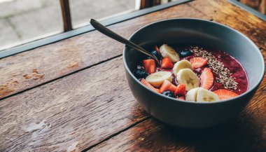 Renkli bir meyve kasesi yıpranmış ahşap bir masada oturur, canlı, sağlıklı bir kahvaltı sahnesi için muz dilimleri, çilekler, kivi ve yabanmersini sergiler..