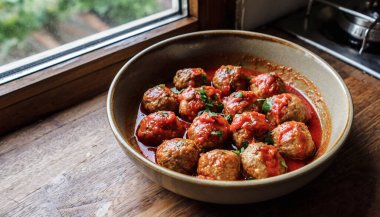 Zengin domates sosuyla pişirilmiş bir kase köfte, ahşap bir pencere kenarında duruyor. Sıcak bir köy yemeği için doğal ışıkta yıkanmış otlarla birlikte.