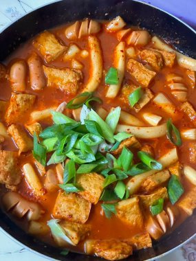 Çiğnenmiş kırmızı soslu pirinç kekleri ve susam tohumları ve yeşil soğanla kaplı baharatlı Kore tteokbokki. Bağımlılık yaratan tadı ve tatmin edici dokusuyla bilinen popüler bir sokak yemeği..