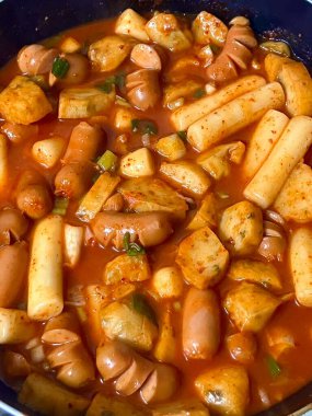 Çiğnenmiş kırmızı soslu pirinç kekleri ve susam tohumları ve yeşil soğanla kaplı baharatlı Kore tteokbokki. Bağımlılık yaratan tadı ve tatmin edici dokusuyla bilinen popüler bir sokak yemeği..