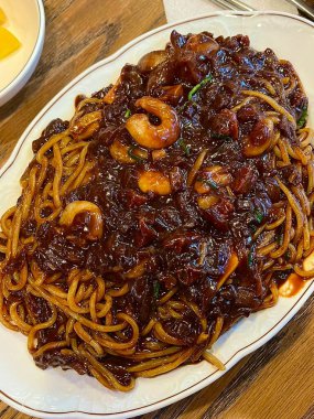 Bol siyah fasulye soslu kalın erişteli klasik jajangmyeon. Dilimlenmiş salatalık ve susam tohumlarıyla süslenmiş. Çok sevilen bir Kore-Çin konfor yemeği, lezzet ve derinlik dolu..