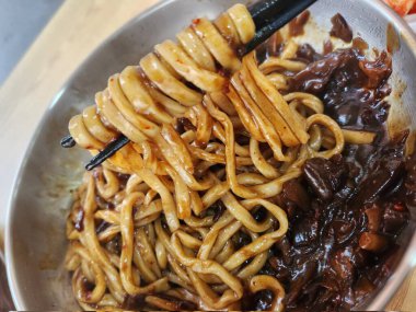 Bol siyah fasulye soslu kalın erişteli klasik jajangmyeon. Dilimlenmiş salatalık ve susam tohumlarıyla süslenmiş. Çok sevilen bir Kore-Çin konfor yemeği, lezzet ve derinlik dolu..