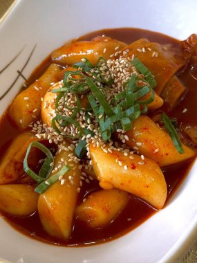 Çiğnenmiş kırmızı soslu pirinç kekleri ve susam tohumları ve yeşil soğanla kaplı baharatlı Kore tteokbokki. Bağımlılık yaratan tadı ve tatmin edici dokusuyla bilinen popüler bir sokak yemeği..