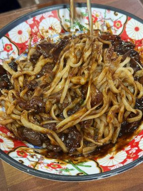 Bol siyah fasulye soslu kalın erişteli klasik jajangmyeon. Dilimlenmiş salatalık ve susam tohumlarıyla süslenmiş. Çok sevilen bir Kore-Çin konfor yemeği, lezzet ve derinlik dolu..