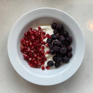 Taze renkli meyvelerle kaplı bir kasede servis edilen Yunan yoğurdu, kremalı pürüzsüz bir doku, canlı doğal renkler ve detaylı bir şekilde ferahlatıcı bir görünüş sunuyor.
