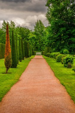 Beautiful English Garden Path with Lush Greenery, Manicured Trees, and a White Bench Under a Cloudy Sky