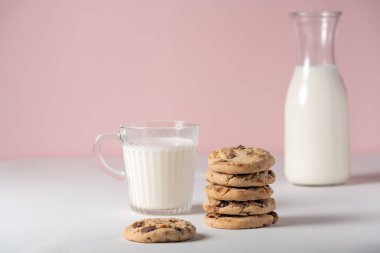 Çerçevenin ortasında ev yapımı çikolata parçacıklı ve fındıklı kurabiyeler. Pembe arka planda bulanık süt kapları var.