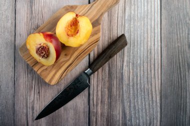 İki canlı nektarin yarım ve küçük, rustik ahşap bir kesme tahtası üzerinde dinlenen profesyonel bir mutfak bıçağı, hafif ahşap yüzey ile çelişiyor.