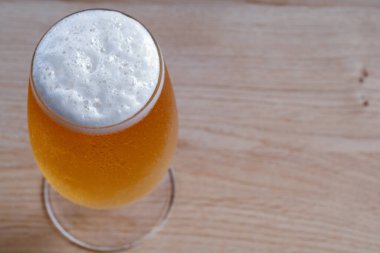 Bir bardak bira ahşap bir masada oturur. Biranın üzerindeki köpük beyaz ve kabarık.