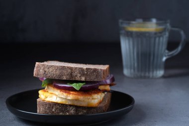 Bir bardak taze limonlu suyla servis edilen, siyah tabakta ızgara halloumi sandviçi içeren dengeli bir yemek. Temiz ve modern gıda tasarımı