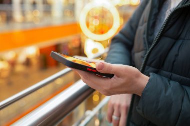 Parlak ışıklı bir alışveriş merkezinde elinde akıllı telefon tutan bir insanın yakın çekimi. Sıcak bokeh ışıkları, modern iletişim, dijital yaşam tarzı, hareketlilik kavramları. 