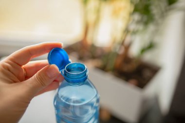 Kadın eli, evdeki bir pencerenin yanında açık plastik bir su şişesi tutuyor. Hidrasyon konsepti, günlük yaşam tarzı, sağlıklı rutin..