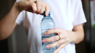Arka planda bulanık çiçekler olan açık bir masadaki plastik şişeyi kapat. Genç bir kadının eli bağlı PET kapağını açar, günlük yaşamı, suyu ve çevre dostu kullanımı vurgular..