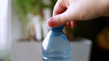 Mutfak masasındaki plastik şişeyi kapat. Arka planda bulanık çiçekler ve pencere olsun. Genç bir kadının eli bağlı PET kapağını açar, günlük kullanım, su ve eko